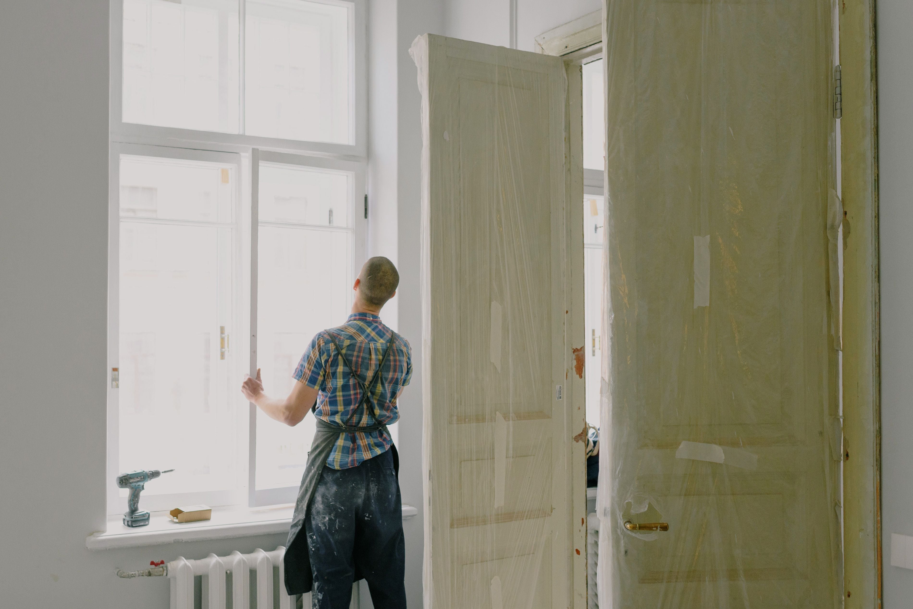 Un artisan devant la fenêtre d'une pièce en rénovation, les portes protégées sous plastique